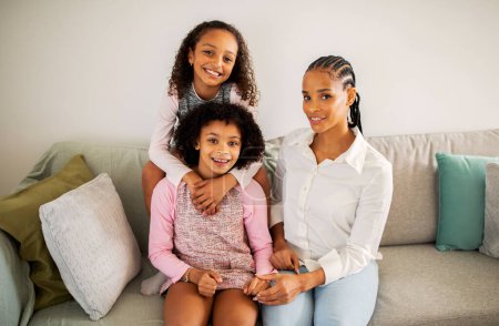 Téléchargez les photos : Maman et filles noires étreignant assis sur le canapé souriant à la caméra à la maison. Famille positive de trois posant ensemble dans le salon moderne. Joyeux concept de maternité et de garde d'enfants - en image libre de droit