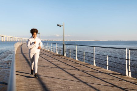 Téléchargez les photos : Mode de vie actif. Heureuse jeune femme afro-américaine en vêtements de sport Jogging en plein air, Athlétique Black Millennial Lady Course sur jetée en bois près de la mer, Profitant de l'entraînement à l'extérieur, Espace de copie - en image libre de droit