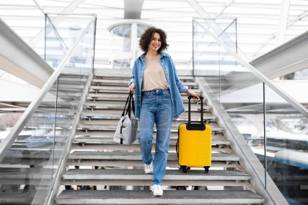 Foto de Sonriente hermosa mujer joven caminando con maletas en las escaleras en el aeropuerto, feliz milenaria viajando sola, llevando equipaje mientras va a la puerta de embarque de vuelo en la terminal, espacio para copias - Imagen libre de derechos