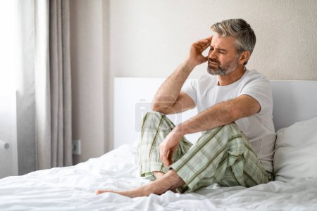 Photo for Sad middle aged handsome grey-haired bearded man in pain sitting in bed, touching his head, suffering from headache, migraine, hangover, wearing pajamas, white bedroom interior, copy space - Royalty Free Image