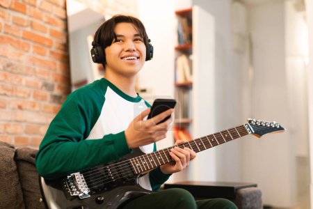 Téléchargez les photos : Talent et Hobby. Joyeux adolescent coréen jouant de la guitare électrique apprenant la musique en ligne tenant Smartphone assis à l'intérieur, portant des écouteurs. Concept d'application technologique et musicale - en image libre de droit