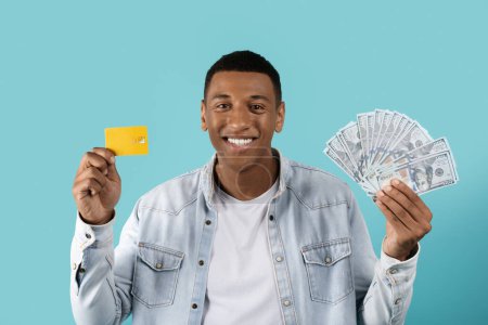 Foto de Satisfecho millennial hombre negro mostrar dólares, tarjeta de crédito, regocijarse con la victoria en línea, recomienda ahorros aislados en el fondo azul. Elección de las finanzas de almacenamiento, dinero de ganancias, reembolso y compras - Imagen libre de derechos