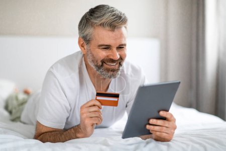 Photo for Positive smiling attractive grey-haired mature man wearing pajamas enjoying online shopping while relaxing at weekend at home, lying on bed, holding digital tablet and bank card, copy space - Royalty Free Image