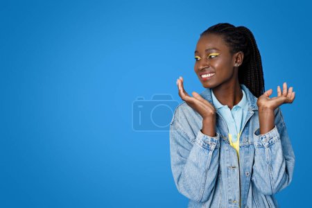Téléchargez les photos : Émotionnel heureux joyeux attrayant jeune afro-américaine dame portant tenue décontractée et lumineux geste de maquillage, tenant les mains à côté du visage, en regardant l'espace de copie pour la publicité sur fond bleu - en image libre de droit