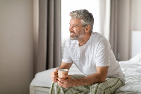 Téléchargez les photos : Joyeux heureux beau barbu aux cheveux gris homme d'âge moyen européen en pyjama assis sur le lit à la maison, tenant une tasse, buvant du café le matin, regardant l'espace de copie et souriant - en image libre de droit