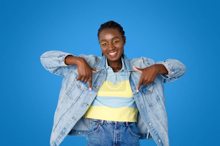 Photo for Smiling cheerful pretty young black woman wearing casual outfit and bright makeup fashionista pointing down with both hands, posing on blue studio background, copy space - Royalty Free Image