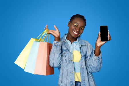Foto de Alegre atractiva joven mujer negra en fashionista casual mostrando compras y teléfono con pantalla vacía en negro, sonriendo, compras femeninas en línea, utilizando la aplicación móvil, fondo azul, maqueta - Imagen libre de derechos