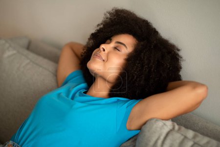 Foto de Alegre señora rizada afroamericana milenaria con los ojos cerrados disfruta de dormir en el sofá en el interior de la sala de estar. Atención médica, descanso y relax en tiempo libre, estilo de vida y descanso - Imagen libre de derechos