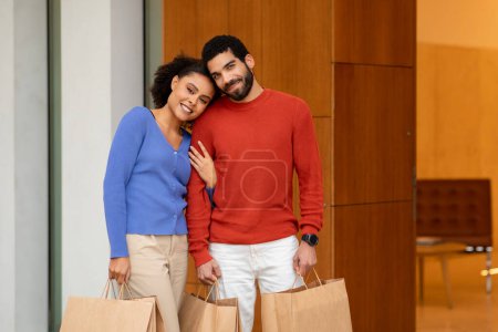 Téléchargez les photos : Joyeux couple multiculturel d'acheteurs faisant du shopping souriant à la caméra debout et étreignant en plein air. Épouses Shopaholics posant avec beaucoup de sacs en papier le week-end. Grand concept d'offre de vente - en image libre de droit
