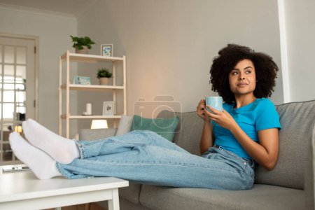 Photo for Thoughtful millennial african american curly lady enjoy relax and cup of tea on sofa, think, look at free space in living room interior. Evening relax alone at home, ad and offer - Royalty Free Image