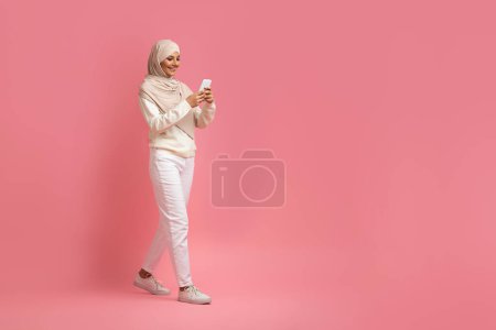 Foto de Comunicación móvil. Mujer musulmana sonriente en Hijab caminando con teléfono inteligente sobre fondo rosa, Mujer islámica feliz navegando por Internet o mensajería con amigos, tiro de cuerpo entero, espacio de copia - Imagen libre de derechos
