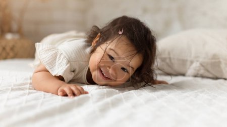 Téléchargez les photos : Adorable bébé coréen bébé fille souriante regardant la caméra allongée sur l'estomac dans la chambre à coucher à la maison. Mignon petit tout-petit enfant rampant sur le lit s'amusant à l'intérieur, portant des vêtements décontractés. Panorama - en image libre de droit