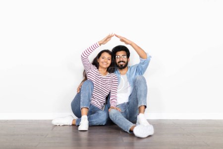 Photo for Happy young mixed race couple sitting on the ground leaning against an exterior white blank wall with copy space, making roof above their heads and smiling. Moving, relocation, mortgage - Royalty Free Image