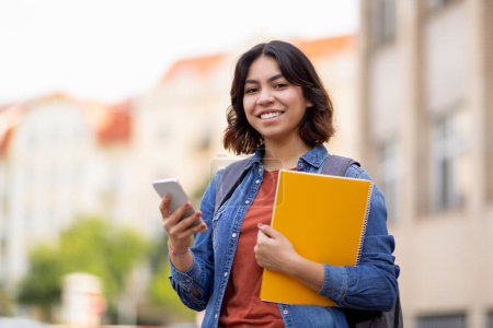 Téléchargez les photos : Portrait extérieur d'une étudiante arabe souriante avec smartphone et cahiers d'exercices, heureuse jeune femme du Moyen-Orient utilisant un téléphone portable tout en se tenant dehors sur le campus, se reposant après les cours - en image libre de droit
