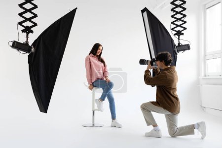 Téléchargez les photos : Photographe professionnel prenant une photo de jeune femme en tenue décontractée, ayant photoshoot dans un studio moderne avec équipement d'éclairage sur fond blanc - en image libre de droit