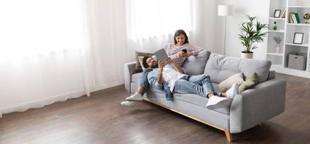 Photo for High angle view of happy smiling multiracial millennial couple relaxing together on couch at home, handsome arab guy wearing glasses using digital tablet lying on indian lady laps, copy space, banner - Royalty Free Image