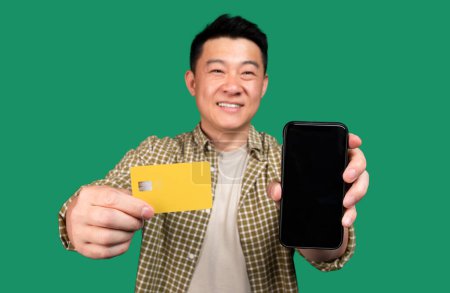 Téléchargez les photos : Application de shopping mobile. Homme coréen joyeux tenant la carte de crédit dans la main et montrant le téléphone portable avec écran vide à l'appareil photo, gadget avec espace de copie pour la maquette, salaire numérique - en image libre de droit