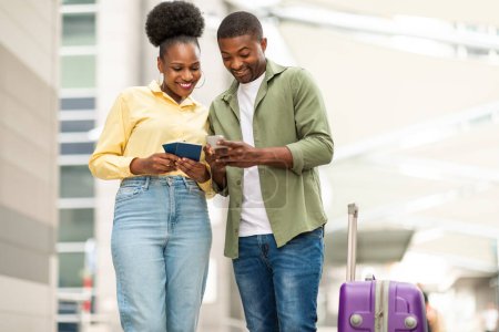Photo for Black Tourists Couple Using Smartphone Holding Passports Booking Travel Tickets In Mobile Application Standing With Suitcase At Airport Outdoor. Technology And Tourism Concept - Royalty Free Image