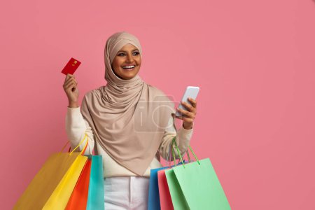 Téléchargez les photos : Souriant dame islamique portant Hijab tenant Smartphone, Carte créadit et sacs à provisions, Heureuse femme musulmane en foulard faisant des achats en ligne tout en se tenant sur fond rose Studio, Copier l'espace - en image libre de droit