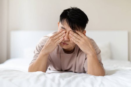 Photo for Closeup photo of sleepy tired adult asian man wearing pajamas lying on bed at home, rubbing his eyes after waking up in the morning, slept bad, feeling powerless. Insomnia, sleeping disorder concept - Royalty Free Image