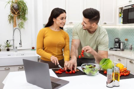 Photo for Newly married couple happy young newlywed making delicious healthy dinner together at home, cutting fresh vegetables, have conversation, watching food blog on Internet, using pc, copy space - Royalty Free Image