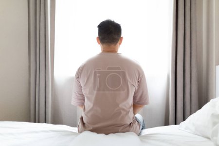 Photo for Back view of unhappy frustrated man wearing pajamas homewear sitting on bed alone at home, suffering from depression, loneliness, anxiety, looking through window, copy space. Mental disorder, illness - Royalty Free Image