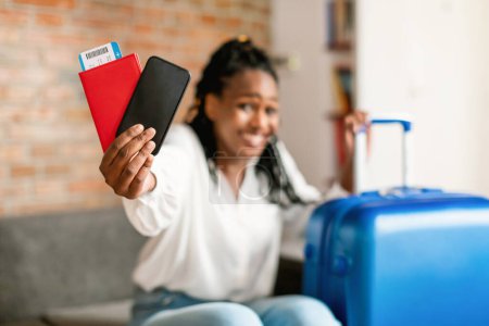 Téléchargez les photos : Heureuse femme afro-américaine montrant un smartphone avec écran vierge et tenant un passeport avec des billets, assise près de valise emballée à la maison, foyer sélectif - en image libre de droit