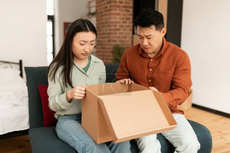 Photo for Disappointed asian married spouses unpacking cardboard box, received damaged item or unwanted stuff, sitting on sofa at home. Annoyed customers dissatisfied by delivery service - Royalty Free Image