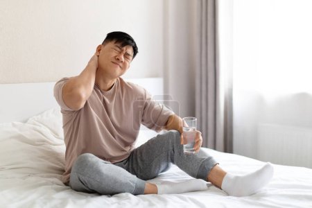 Foto de Infeliz madura asiática hombre en dolor sentado en la cama en casa, frotando su cuello, sufren de tensión muscular de la mañana después de dormir en la cama incómoda, sosteniendo un vaso de agua, tomando analgésicos, espacio de copia - Imagen libre de derechos