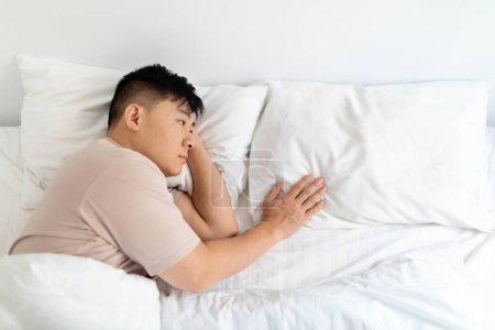Téléchargez les photos : Solitaire malheureux moyen homme asiatique veuf allongé dans le lit seul, se sentant triste, touchant et regardant oreiller vide à côté de lui, passant par la rupture ou le divorce, intérieur de la maison, espace de copie - en image libre de droit