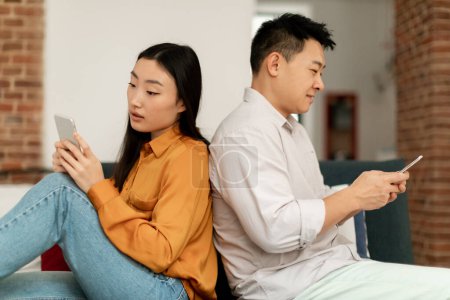 Foto de Crisis conyugal, indiferencia. Esposos coreanos usando teléfonos celulares, sentados espalda con espalda en el sofá, indiferentes el uno al otro, vista lateral. Pareja ignorando unos a otros, mensajes de texto con los amantes - Imagen libre de derechos