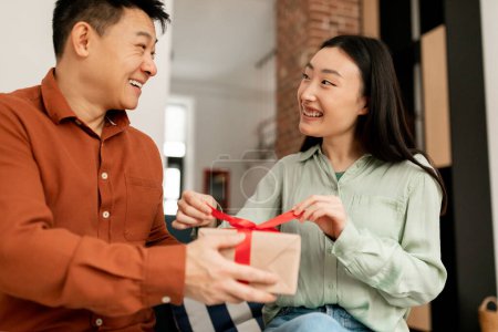Photo for Anniversary and birthday celebration concept. Happy korean woman opening gift with red ribbon from her husband, sitting on sofa in living room interior, looking at each other and smiling - Royalty Free Image