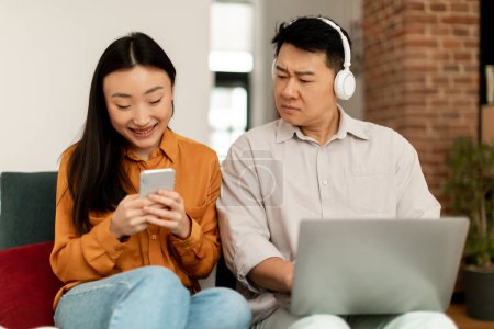 Photo for Marital problem. Jealous asian middle aged man looking at wifes smartphone while working on laptop and wearing headphones, sitting together on sofa - Royalty Free Image