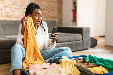 Photo for Terrified black lady reading shocking travel news on smartphone while packing suitcase at home in living room. Cancelled flight, traveling problems concept - Royalty Free Image