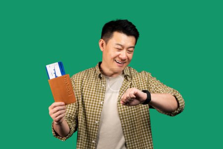 Téléchargez les photos : Heureux homme asiatique tenant un passeport avec des billets, regardant la montre et souriant, vérifiant l'heure sur fond vert, studio tourné. Voyage, tourisme, concept de ponctualité - en image libre de droit