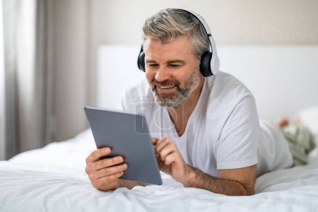 Photo for Relaxed smiling handsome grey-haired bearded middle aged man wearing pajamas lying on bed at home, using digital tablet and wireless headset in bed, watching TV show online, free space - Royalty Free Image