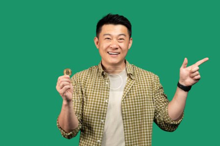 Foto de Feliz asiático de mediana edad hombre sosteniendo bitcoin moneda virtual y señalando a un lado en el espacio libre, mirando y sonriendo a la cámara, de pie sobre fondo de estudio verde - Imagen libre de derechos