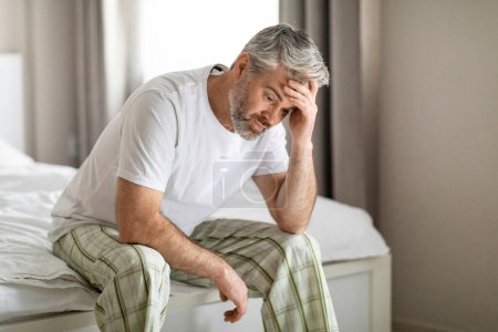 Photo for Upset thoughtful middle aged grey-haired handsome man wearing pajamas sitting on bed alone at home, touching head, suffering from hangover or anxiety, copy space. Permacrisis, midlife crisis - Royalty Free Image