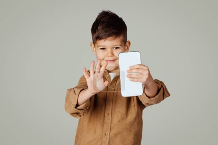 Photo for Cheerful caucasian 6 years old little boy waving hand on smartphone, has video call isolated on gray studio background. App for communication with device, lifestyle and lesson remotely - Royalty Free Image
