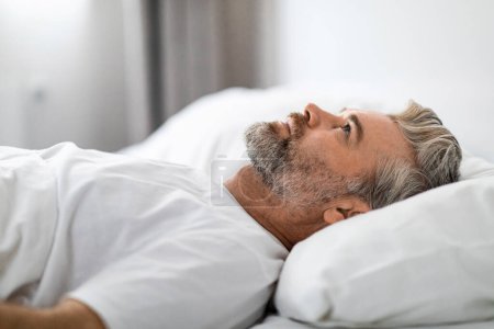 Photo for Side view of unhappy frustrated handsome grey-haired bearded middle aged man wearing pajamas lying in bed alone, looking at ceiling, male suffering from depression, closeup - Royalty Free Image