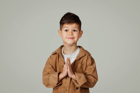 Téléchargez les photos : Joyeux petit garçon caucasien mignon de 6 ans dans un geste occasionnel avec les mains priant isolé sur fond de studio gris. Demander, rêve devenu réalité, mode de vie de l'enfance, étude et expression faciale - en image libre de droit