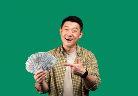Photo for Happy korean middle aged man holding dollar cash money and pointing at it, recommending fast credit and smiling, standing over green studio background - Royalty Free Image