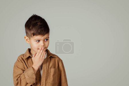 Photo for Smiling shocked cute caucasian 6 years old little boy in casual closes mouth with hand look at free space isolated on gray studio background. Fun, childhood, education ad and offer, lifestyle - Royalty Free Image