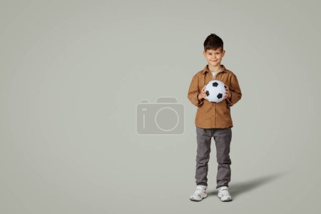 Téléchargez les photos : Souriant caucasien de 6 ans petit garçon décontracté avec ballon de football, profiter du jeu, isolé sur fond de studio gris. Avenir avec le sport, passe-temps et jeu, émotions de l'enfant mode de vie de l'enfance - en image libre de droit