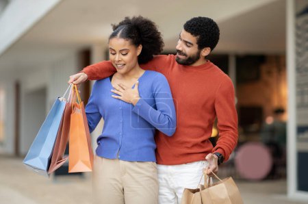 Photo for Family Shopping. Couple Enjoying Weekend In Modern Mall, Cheerful Boyfriend Surprising Girlfriend Giving Paper Shopper Bags To Her Standing Outside. Commerce And Consumerism - Royalty Free Image
