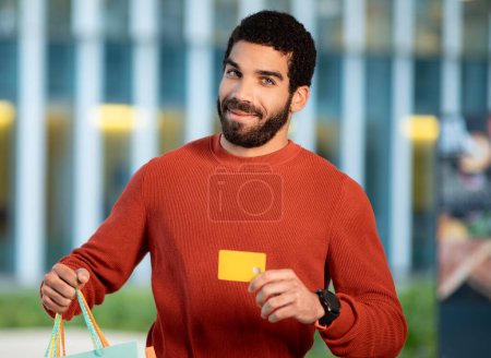 Foto de Hombre de Oriente Medio mostrando la tarjeta de crédito sonriendo a la cámara sosteniendo bolsas de compras de papel, la publicidad de su banco de pie al aire libre en el área urbana. Hombre comprador aprobación gran oferta financiera - Imagen libre de derechos