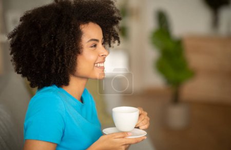 Téléchargez les photos : Joyeux jeune femme frisée afro-américaine profiter de la détente et une tasse de thé sur le canapé dans le salon intérieur, floue, profil. Soirée détente seule, temps libre le matin, annonce de café et offre - en image libre de droit