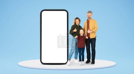 Photo for Cheerful adult caucasian family show thumbs up, approving ad and offer on huge smartphone with blank screen on blue studio background. Like, app, website for family communication - Royalty Free Image