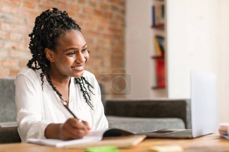 Téléchargez les photos : Femme d'affaires noire assise à table et prenant des notes dans un carnet, assistant à un webinaire ou à une formation via un ordinateur portable à la maison. Apprentissage féminin en ligne, participation à un cours éducatif, espace de copie - en image libre de droit