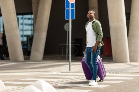 Téléchargez les photos : African American Male Tourist Walking Through Crosswalk Posing With Travel Suitcase Arrivée à l'aéroport moderne en plein air. Prise de vue pleine longueur d'un passager masculin à l'extérieur de la gare - en image libre de droit